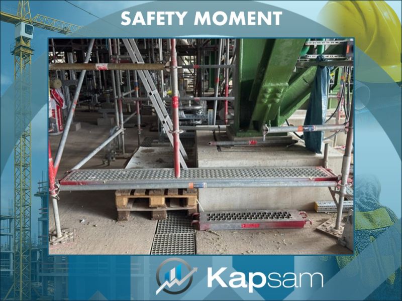"Poster mit der Überschrift 'Safety Moment'. Abgebildet ist ein Baugerüst mit Metallböden und rotem Gerüstgestänge. Ein Gerüststeg liegt unsicher auf Holzpaletten und über einer Öffnung im Boden, wodurch eine Stolper- und Absturzgefahr entsteht. Unten ist das Logo von Kapsam sichtbar."